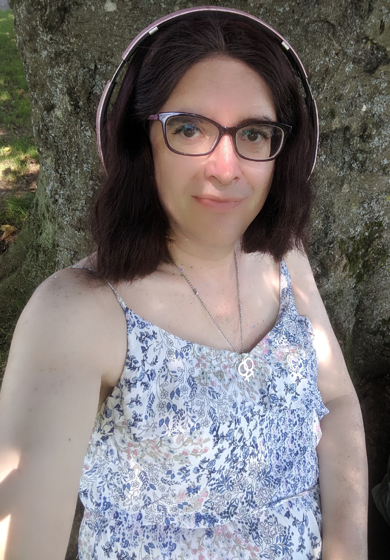 Selfie of me looking at the camera while I'm sat against a tree. I have shoulder length red-tinted black hair cut in a bob, and I have a red fabric hairband holding my hair back off my face. Over the top of the hairband I am wearing my pink headphones