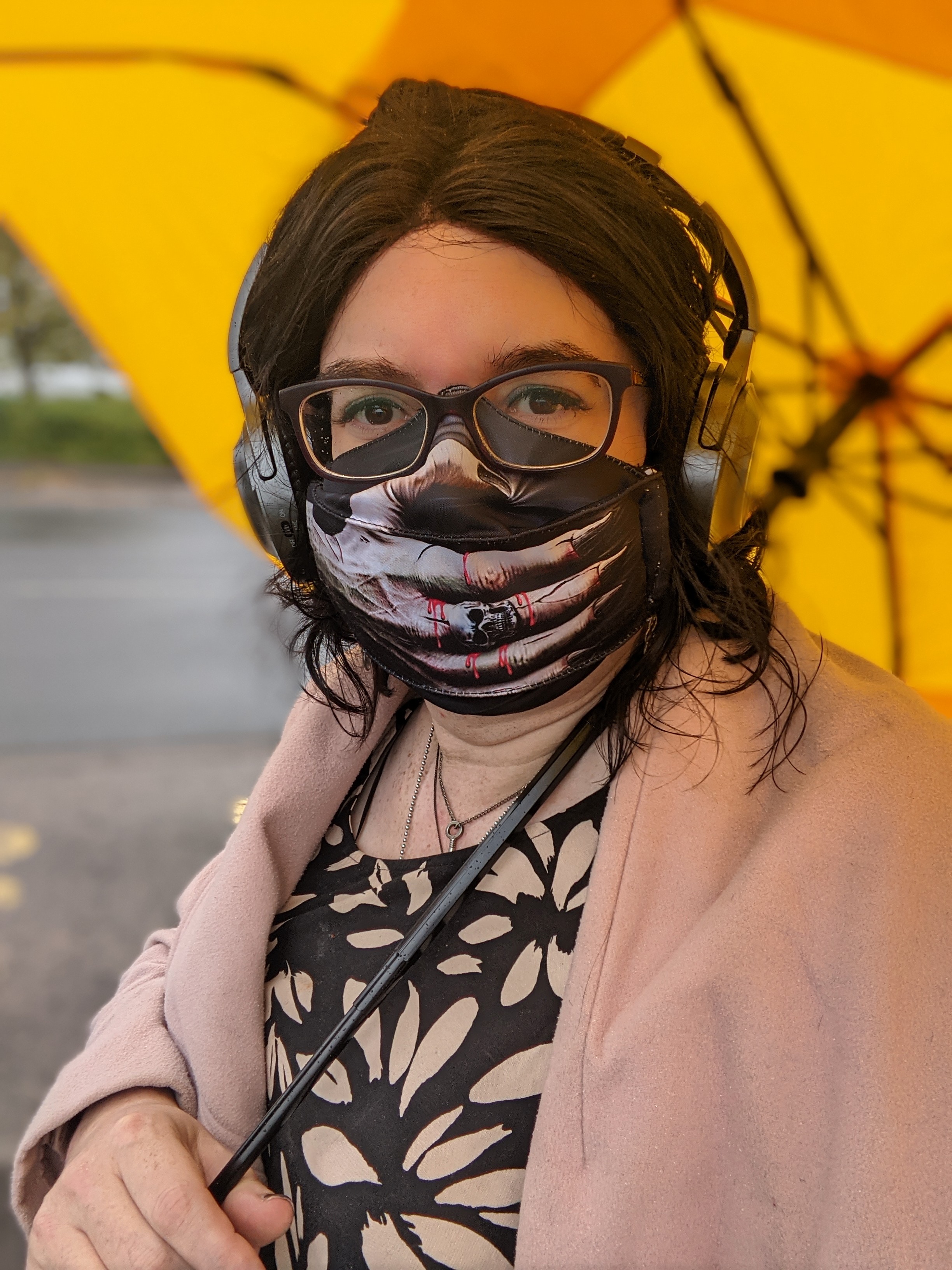 A photo of me. I have shoulder length hair held back off my face with a fabric hairband. I'm wearing a facemask that has an image of a skeletal hand on it stretching over my nose and mouth. I'm holding a yellow umbrella over myself
